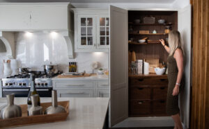 woman with blonde hair reaching into a cupboard which has been designed by colour house interiors in a white kitchen with a large cooker